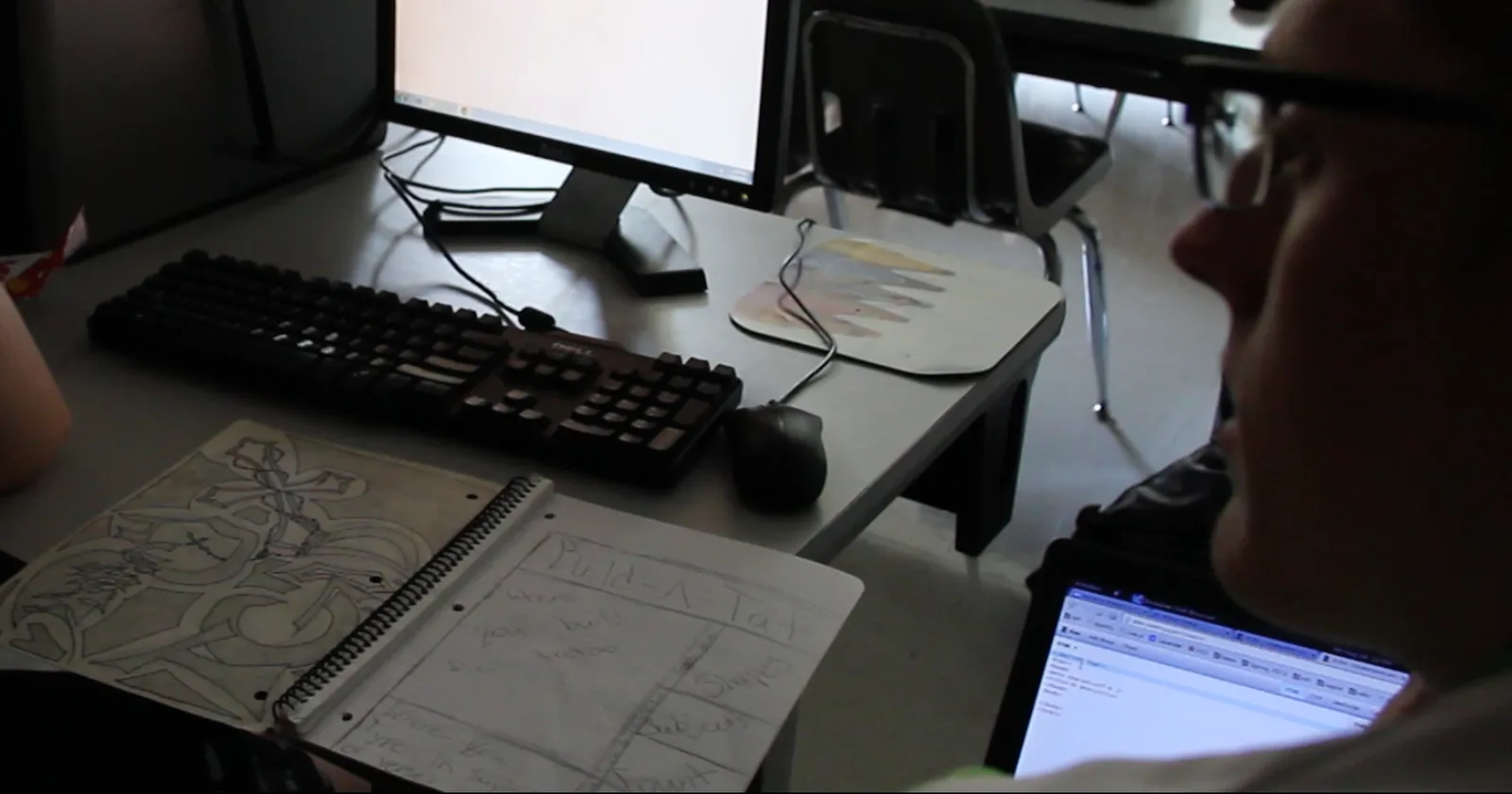 Foto meja komputer di ruang kelas: buku tulis spiral terbuka berisi sketsa wireframe pensil di samping keyboard, mouse, dan monitor, dengan seorang pembimbing di sisi kanan