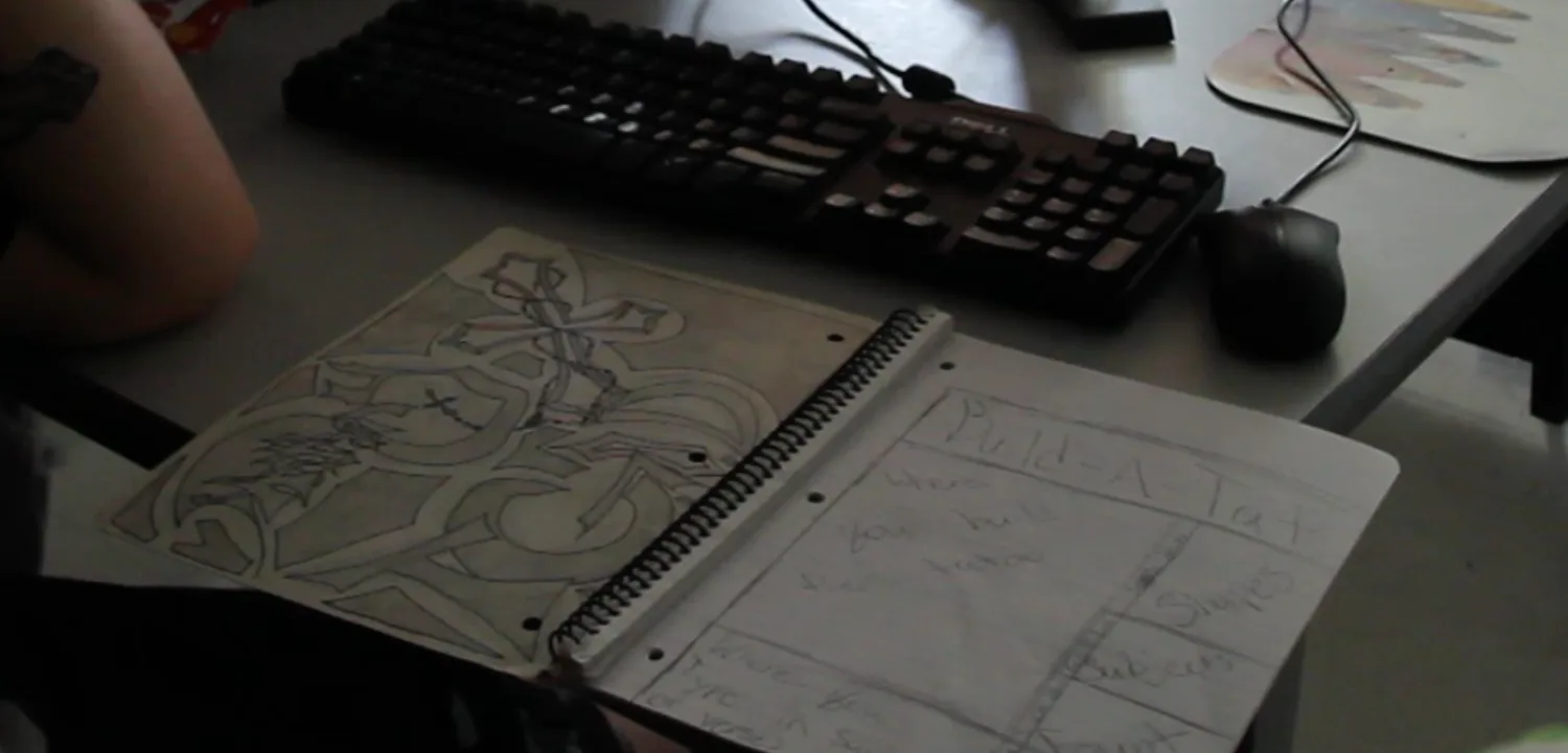 Foto buku tulis spiral terbuka di meja komputer: halaman kanan berisi sketsa wireframe pensil berjudul Build-A-Tat dengan kotak-kotak bertulisan tangan, di belakangnya keyboard dan mouse